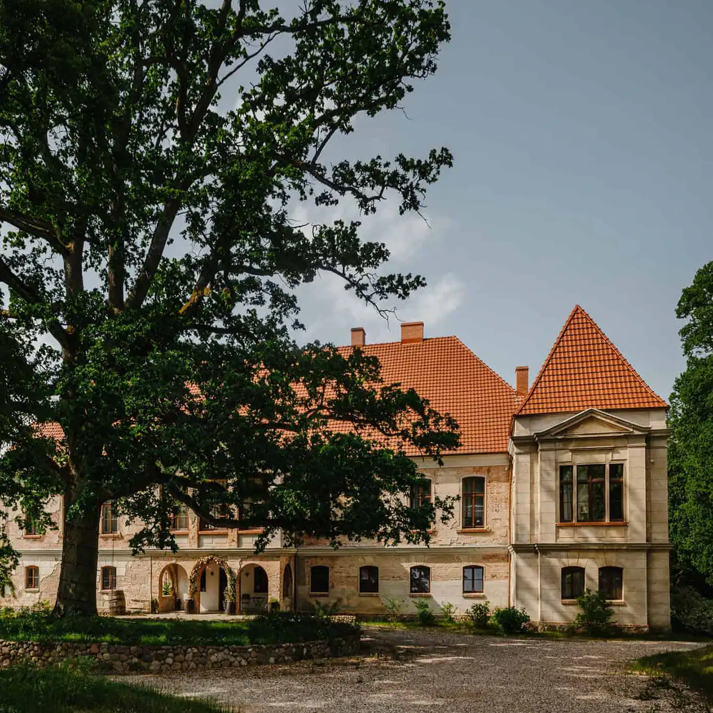 Stary pałac z czerwonym dachem i dużym drzewem w tle.